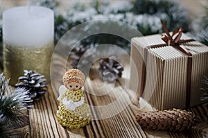 A gift lays on a wooden table next to a candle, cones and an angel against the background of Christmas decorations