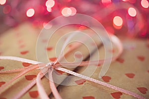 Gift in paper packaging with a pattern of red hearts, ribbon bow and pink lights on background