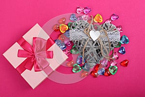 Gift in a pink box on crimson background and wicker decorative heart beside. Gift box with wicker heart beside surrounded with