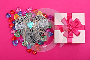 Gift in a pink box on crimson background and wicker decorative heart beside. Gift box with wicker heart beside surrounded with