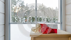 A gift in a red-white box and two hearts on a wooden table near the window with a view of the winter garden