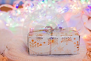 A gift wrapped in a festive packaging tied with a silver ribbon on a knitted white product on a background of bokeh of different-
