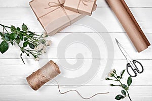 Gift wrapping background. Rolls of kraft wrapping paper, rope, branch of roses, gift boxes and scissors on wooden background
