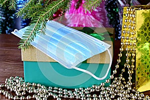 Gifts in beautiful package, in box with green ribbon and gold beads under Christmas tree. On top is disposable medical mask