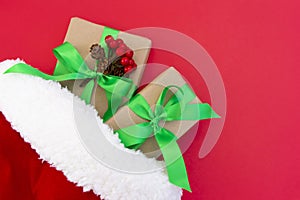 Gifts wrapped in craft paper and decorated with green and red ribbons flat lay on red background next to a Christmas hat