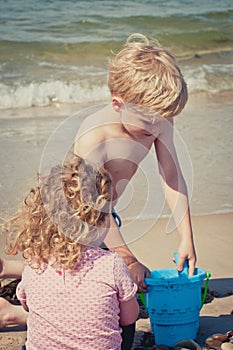 Giirl watches her brother exploring and playing in the sand with a blue bucket