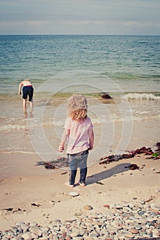 Giirl watches her brother exploring and playing in the sand with a blue bucket