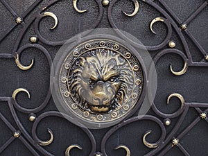 Gilded textured image of a lion`s head with forged ornament on a black iron surface