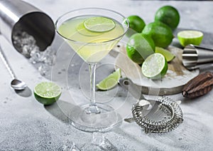 Gimlet Kamikaze cocktail in martini glass with lime slice and ice on light board with fresh limes and strainer with shaker.