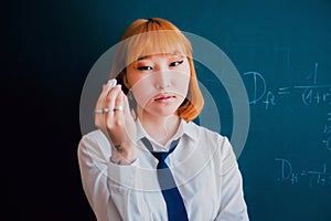 A ginger Asian student, dressed in a uniform, standing against a backdrop of a chalkboard covered with economic formulas. She