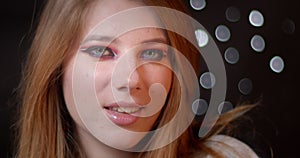 Ginger model with extraordinary bright make-up smiles seductively into camera on blurred lights bokeh background.