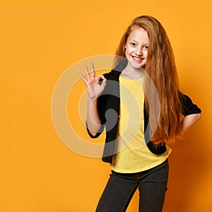 Ginger teenage girl in black jacket, pants, yellow t-shirt. She smiling, showing okay sign, posing on orange background. Close up