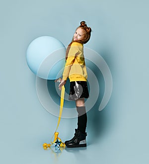 Ginger teenage girl in yellow sweatshirt, black skirt, knee-highs, boots. She smiling, holding balloon, posing on blue background.