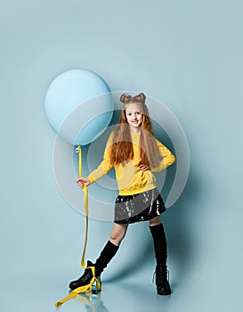 Ginger teenage girl in yellow sweatshirt, black skirt, knee-highs, boots. She smiling, holding balloon, posing on blue background.