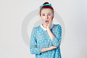 Ginger young girl screaming with shock, holding hands on her cheeks. Isolated studio shot on gray background