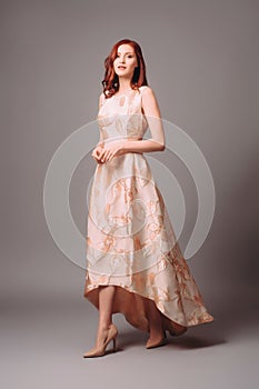 Ginger young woman in golden peach dress with floral print. Calm studio portrait of young lady in long sleeveless evening gown