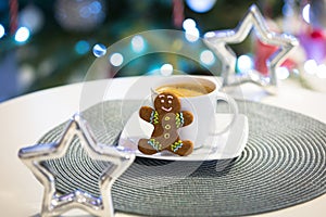 Gingerbread cookie man at the cup of hot coffee and christmas tree lights in background