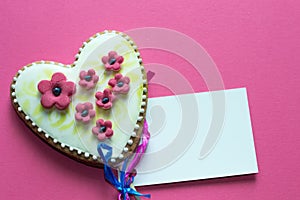 Gingerbread heart with white icing and pink flowers shit of paper on pink background. Love concept. Gift for womans and valentines