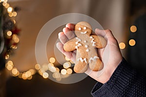 Gingerbread man cookie with sugar icing in hand, blurred garland lights on background. Christmas holiday at home