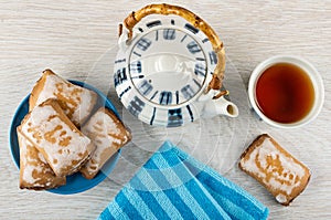Gingerbread in sugar glaze in saucer, blue napkin, teapot, cup of tea on wooden table. Top view