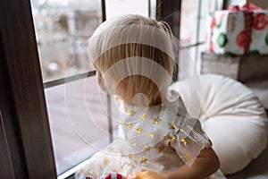 Girl 2 years old sits on the windowsill. A little blonde in a white dress looks out the window.