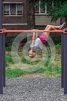 Girl of 6-7 years old is playing cheerfully on the playground, next to the house, and hangs upside down on the uneven bars