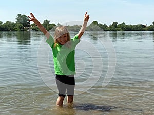 Girl 6 - 7 years old plays with water in the river. The child is wearing a green T-shirt and black shorts. Summer on the pond.