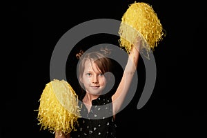 A girl of 6 years, on a black background. A cheerleader with yellow pumpons.Sport and energy.