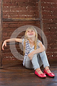 Girl 6 years old in jeans and a vest sits on the floor next to brick wall. filled with laughter