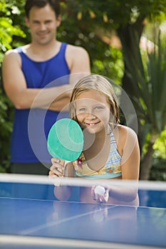 Girl (7-9) playing table tennis with father in background.