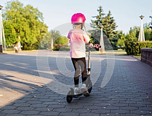 A girl 7 years old in a helmet and a pink injury protection kit rides a scooter in the park, sunny day. Active sports