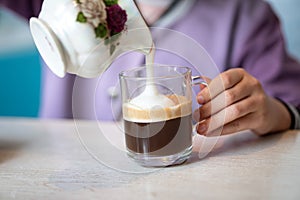 girl adding milk into glass of delicious coffee with whipped cream