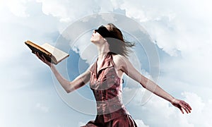 Girl against cloudy sky with opened book in palm as idea for knowledge