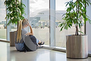 Girl at the airport near window is waiting for a flight with luggage backpack and looking at a plane