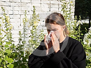 A girl with allergies sneezes into a handkerchief on the street among flowers, the concept of allergic reactions