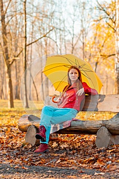 Girl alone sits under an umbrella in an autumn park on a bench cut from a log