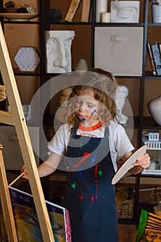 A girl artist in an apron in a creative workshop draws on an easel with paints and a brush. The child is covered in paint