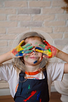 A girl artist in a creative workshop covers her face with her hands stained with paint on the background of a brick wall.