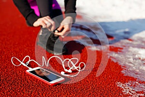 Girl athlete in black sneakers, crouching on the red track for running. near is a phone with wired headphones. Cold snowy weather