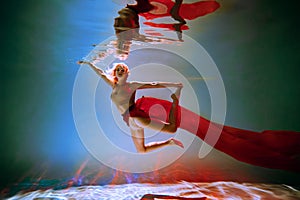 girl with an athletic figure and blond hair, with red material, in a ballet pose underwater