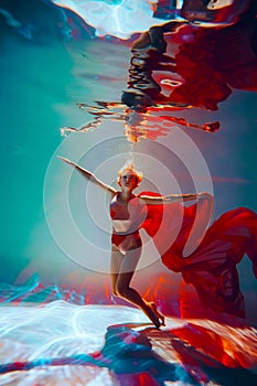 girl with an athletic figure and blond hair, with red material, in a ballet pose underwater