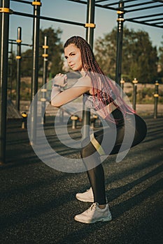 A girl of an athletic physique does an exercise squat with a rubber band expander on a street playground