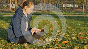 Girl autumn in the park sits on the grass, sending sms from the phone.