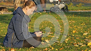 Girl autumn in the park sits on the grass, sending sms from the phone.