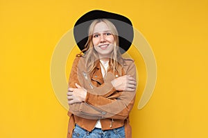 Girl in autumn warm clothes hugs herself with hands and smiles on a yellow isolated background, dress warmly concept