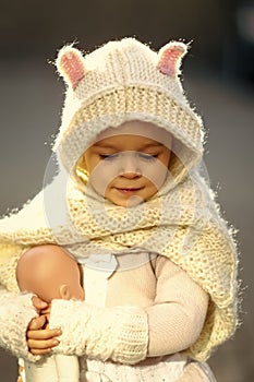 Girl baby in knitted hood and scarf playing with doll