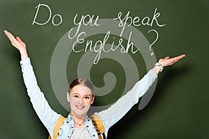 Girl with backpack pointing at chalkboard with do you speak English lettering