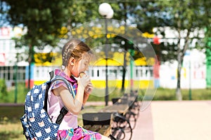 Girl with a backpack sitting on a bench and eating a pie near the school. A quick snack with a bun, unhealthy food, lunch from