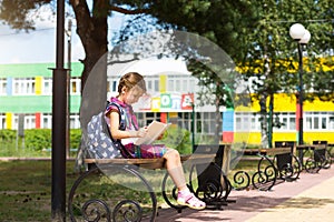 Girl with a backpack sitting on a bench and reading a book near the school. Back to school, lesson schedule, a diary with grades.