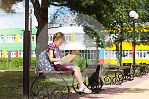 Girl with a backpack sitting on a bench and reading a book near the school. Back to school, lesson schedule, a diary with grades.